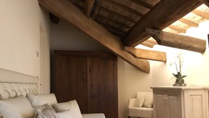 Um quarto aconchegante com teto de madeira, um sofá com almofadas estampadas, uma poltrona branca e um armário com uma planta em vaso.