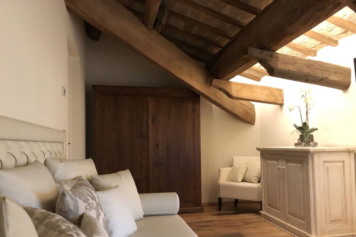 Una habitación acogedora con techo de madera, un sofá con cojines estampados, un sillón blanco y un aparador con planta en maceta.
