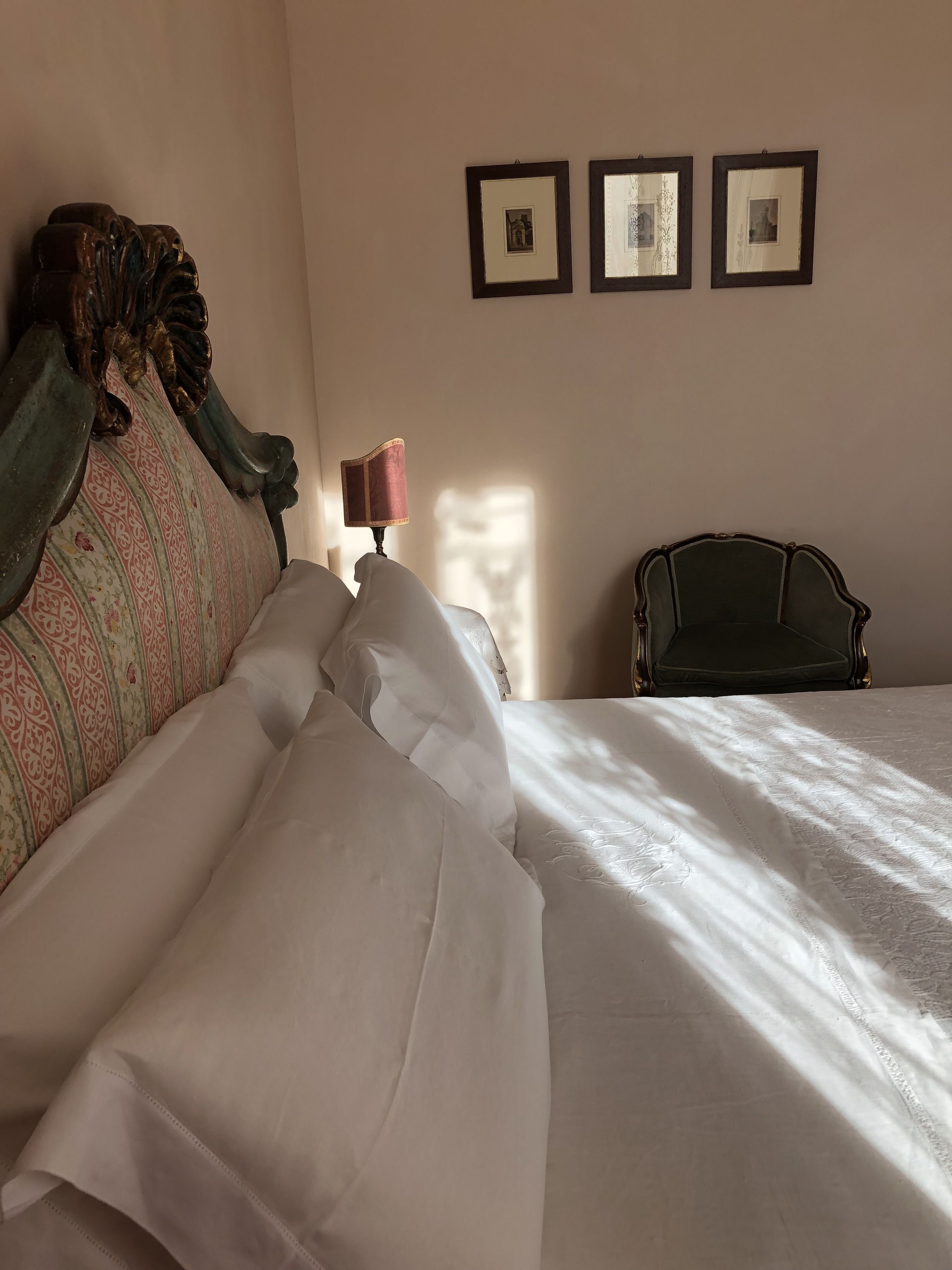 A neatly made bed with white linens, a vintage-style headboard, and a lamp on a bedside table. There are three framed pictures on the wall.