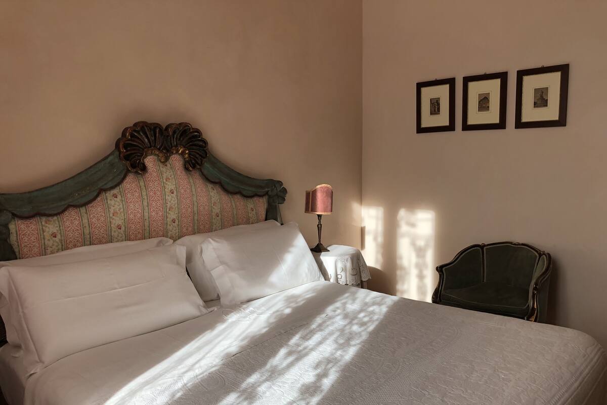 Una cama bien hecha con ropa blanca, un cabecero de estilo antiguo y una mesita de noche con una lámpara. Tres cuadros enmarcados cuelgan de la pared.