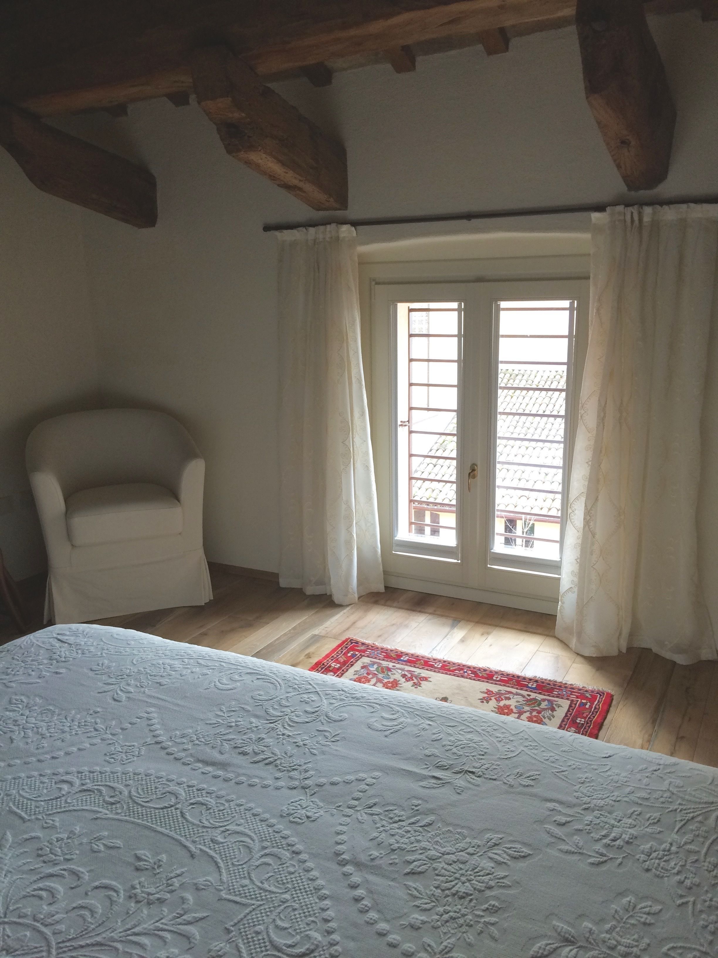 A room with a bed, a chair, a window with curtains, and a wooden ceiling.
