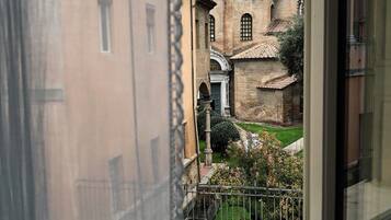 A view through a window of a building with a brick facade and arched windows, a small courtyard with greenery, and a metal railing.