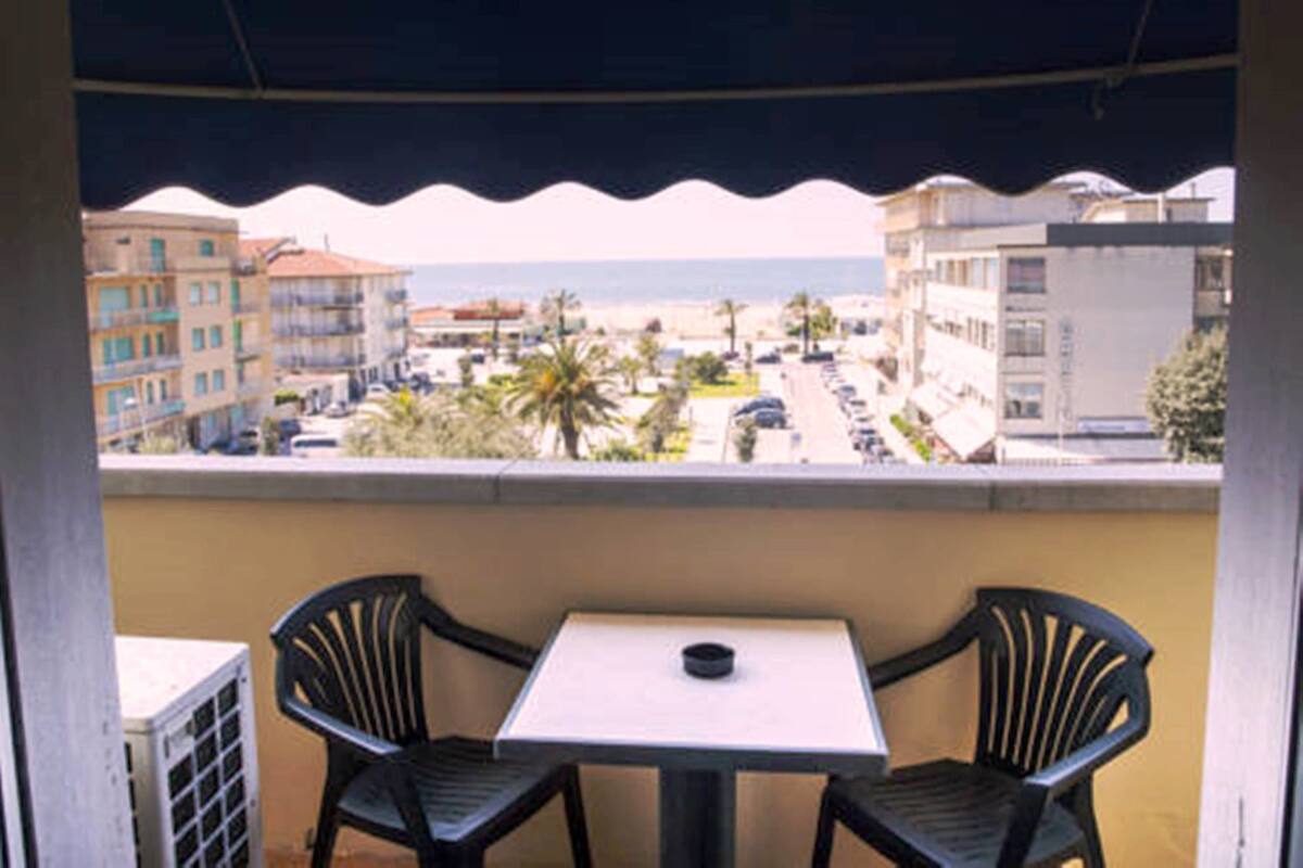 Ein Balkon mit Tisch und zwei Stühlen, mit Blick auf einen Strand und Gebäude.