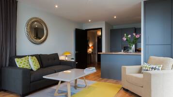 Una sala de estar moderna con un sofá, una mesa de centro y un sillón. Se ve un área de cocina con armarios y una encimera a través de una puerta abierta.