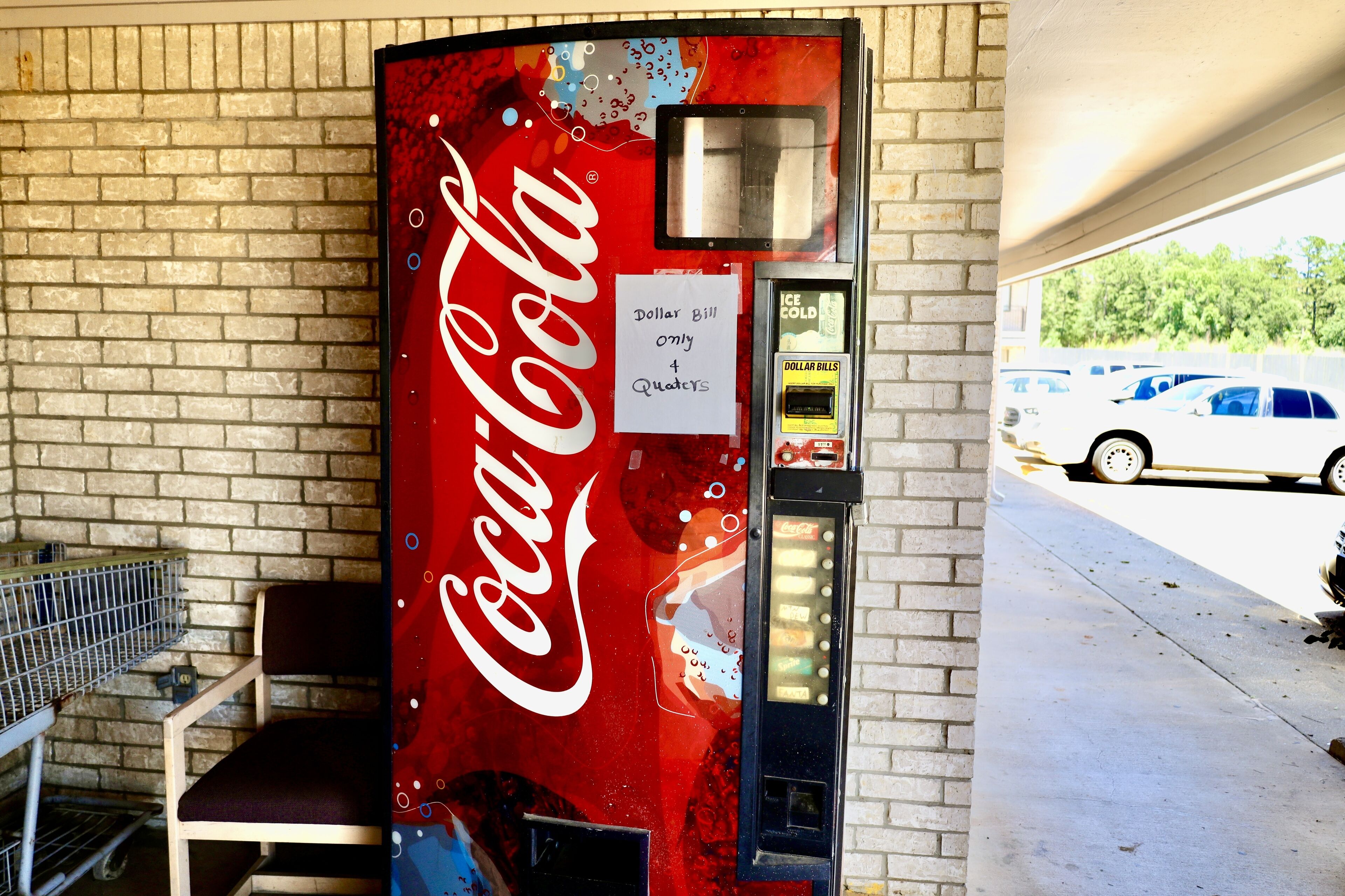 Máquina expendedora de Coca-Cola con un cartel que indica que solo acepta billetes de un dólar o cuatro monedas de veinticinco centavos, ubicada junto a un muro de ladrillo y una zona de estacionamiento.