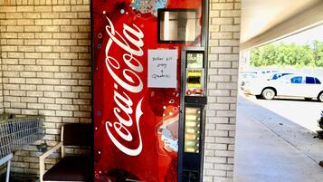 Máquina expendedora de Coca-Cola con un cartel que indica que solo acepta billetes de un dólar o cuatro monedas de veinticinco centavos, ubicada junto a un muro de ladrillo y una zona de estacionamiento.