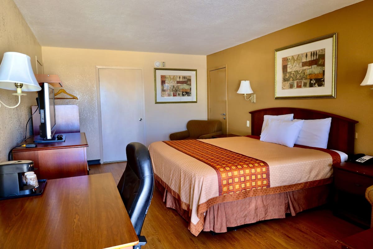 A hotel room with a bed, desk, chair, and a small table with a lamp.