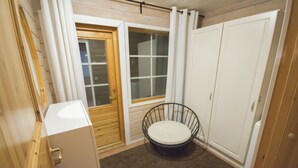 Una habitación pequeña con una puerta de madera, una ventana con cortinas, un armario blanco y un sillón redondo con cojín.