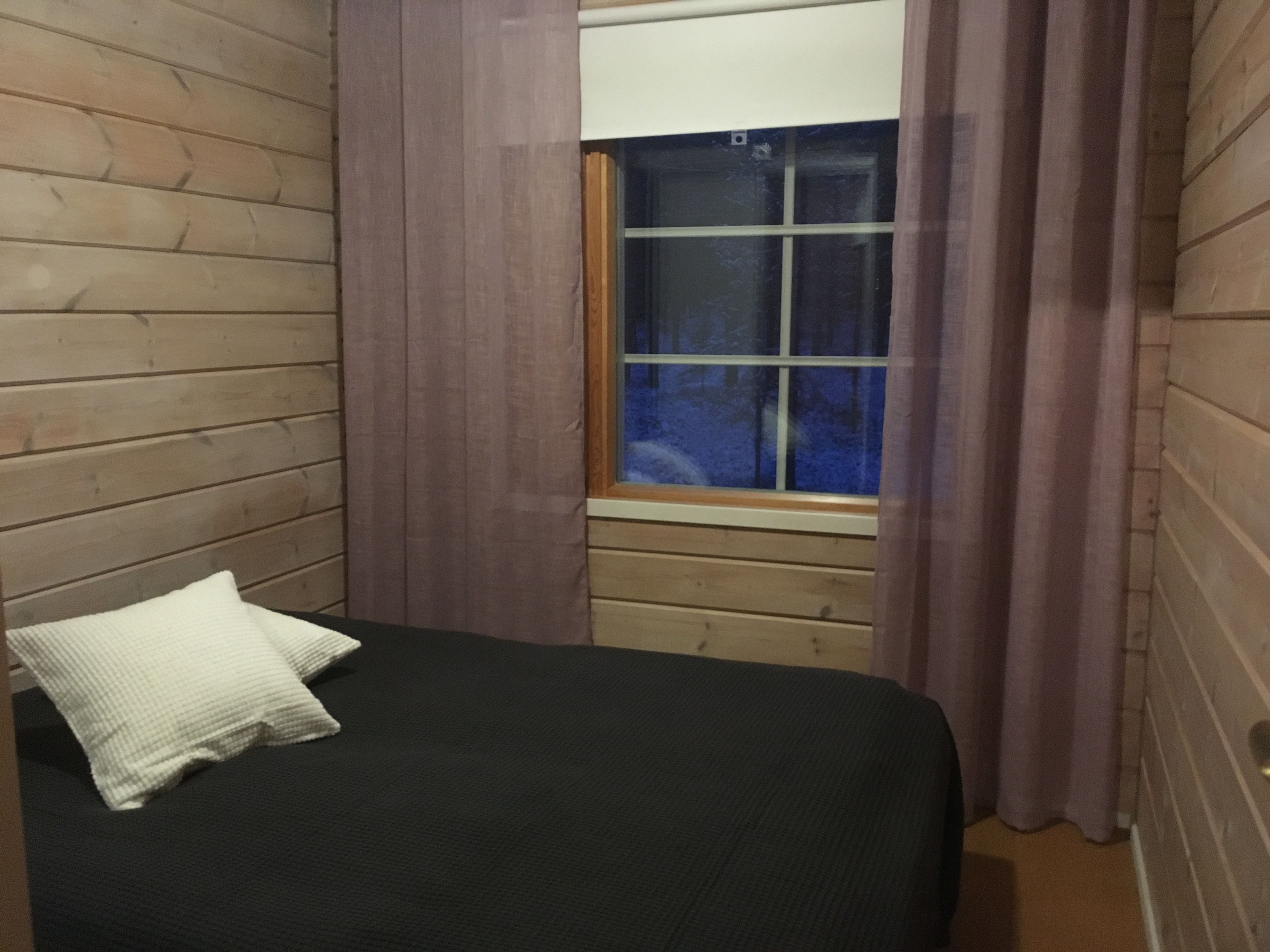 Una habitación con paredes de madera, una ventana con cortinas y una cama con almohada blanca, con vista a árboles.