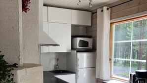 Una cocina compacta con armarios blancos, microondas integrado, refrigerador y ventana con vista a árboles.