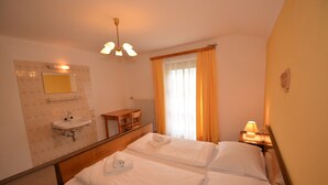 Habitación de hotel con cama, mesita de noche con lámpara, pequeña mesa con silla, espejo y ventana con cortinas.