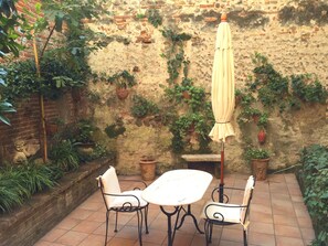 Une terrasse extérieure avec une table et deux chaises, entourée de plantes en pot et d’un mur de pierre.