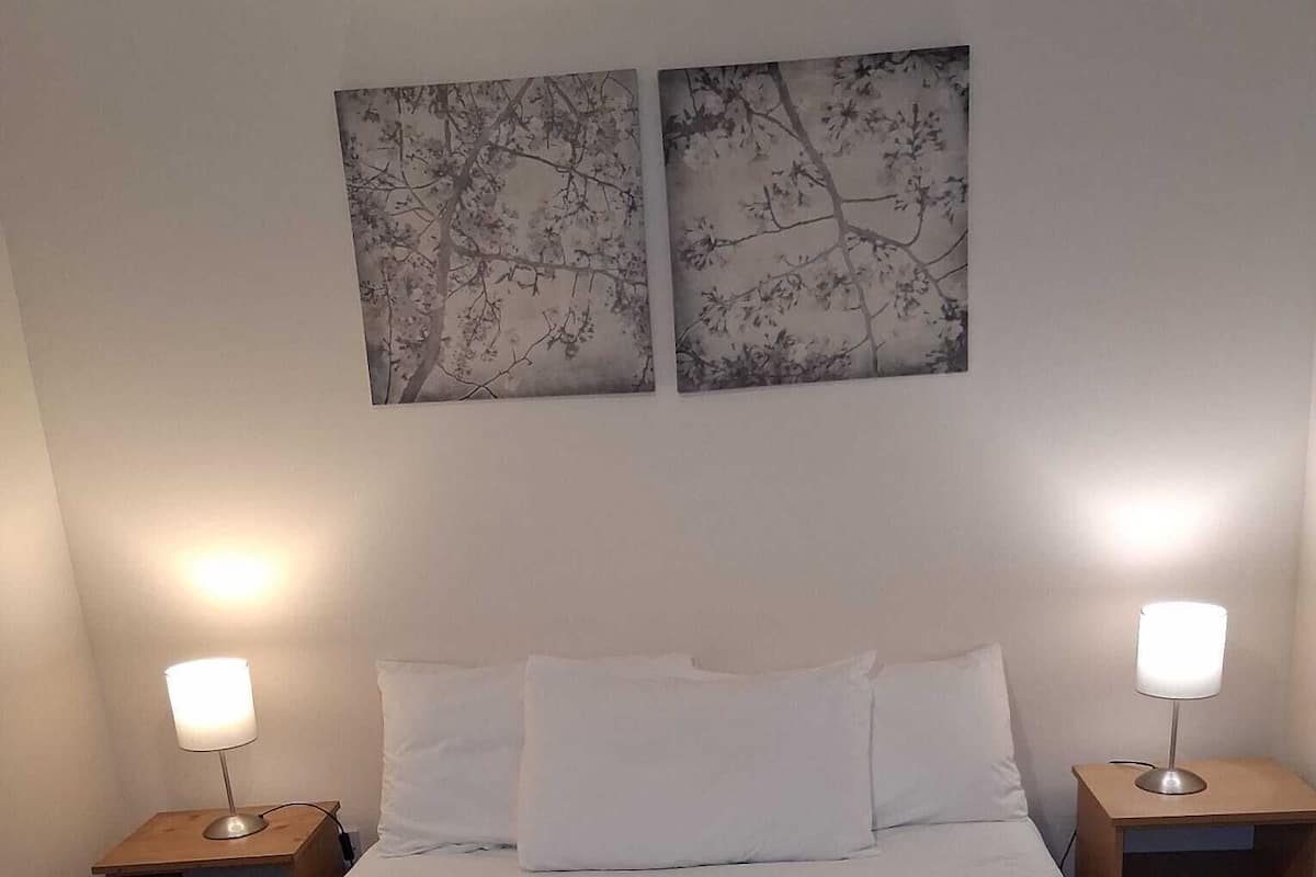 A neatly made bed with white linens and a gray quilt, flanked by two bedside tables with lamps, in a room with plain walls and two framed artworks above the bed.