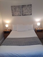 Una cama bien hecha con sábanas blancas y colcha gris, flanqueada por dos mesitas de noche con lámparas, en una habitación con paredes lisas y dos obras de arte enmarcadas sobre la cama.