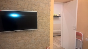 A room with a stone wall, a flat-screen TV mounted on it, a white door, a small shelf with items, and a red chair. - B&B Anny (Naples)