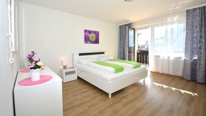 Ein modernes Schlafzimmer mit einem weißen Bett, einem Nachttisch mit Blumenvase, einem Bild an der Wand und Blick ins Freie durch große Fenster.