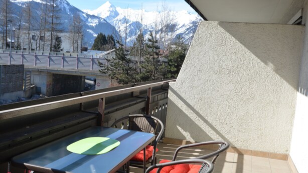 Ein Balkon mit Tisch und Stühlen, mit Blick auf schneebedeckte Berge und eine Brücke.