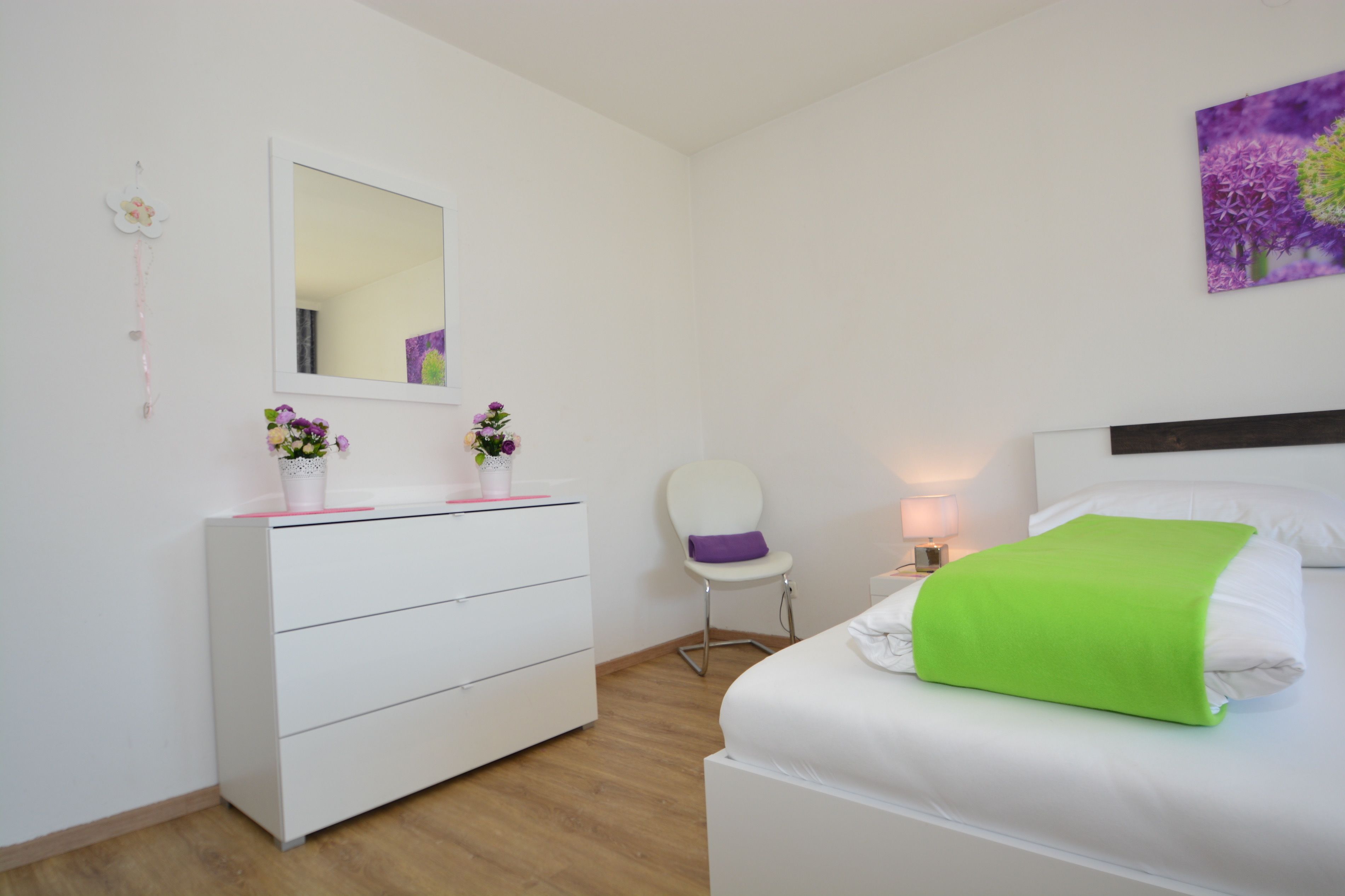 Un dormitorio con un tocador blanco, una silla, una cama con almohada verde y un espejo montado en la pared.