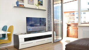 Ein modernes Wohnzimmer mit einem Flachbildfernseher auf einer weißen Unterhaltungseinheit, einer Schiebetür, die auf einen Balkon führt, und Blick auf ein Gebäude.