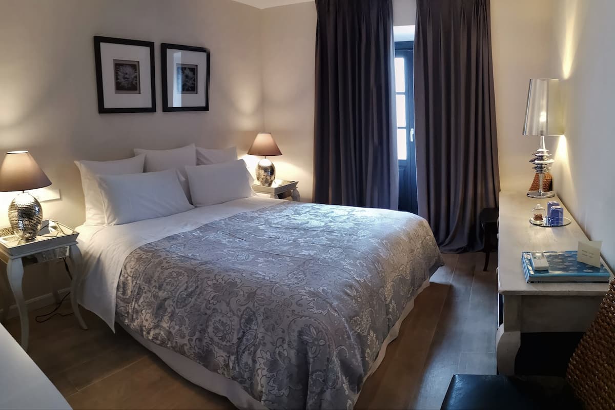 Een keurig opgeruimde hotelkamer met een groot bed, twee bedlampjes, een klein tafeltje met een lamp, en uitzicht op het landschap door een raam met gordijnen.