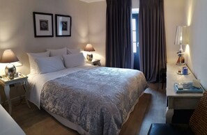 Een keurig opgeruimde hotelkamer met een groot bed, twee bedlampjes, een klein tafeltje met een lamp, en uitzicht op het landschap door een raam met gordijnen.