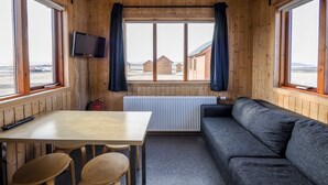 Une cabane en bois comprenant une table à manger, deux chaises, un canapé, un téléviseur fixé au mur et de grandes fenêtres avec des rideaux.