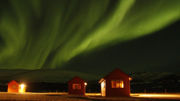 Trois petites cabanes illuminées, se détachant sur le fond des aurores boréales.