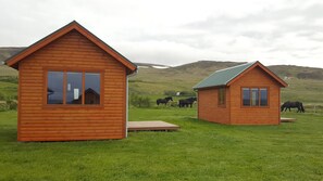 Deux cabanes en bois aux toits en pente, situées dans un champ verdoyant, avec des chevaux en arrière-plan.