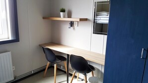 Une petite pièce avec un bureau en bois, deux chaises noires, une plante en pot posée sur une étagère et, au fond, un lit superposé.