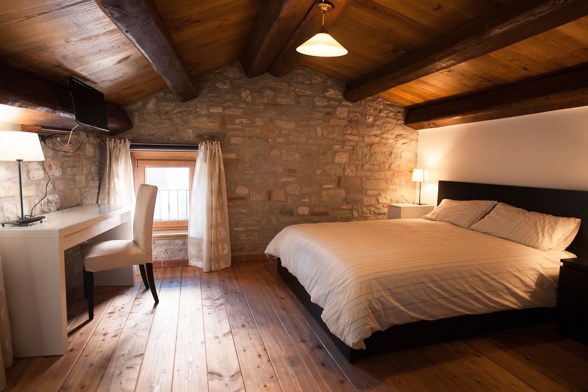A bedroom with a stone wall, wooden ceiling beams, a bed, a desk, and a chair.