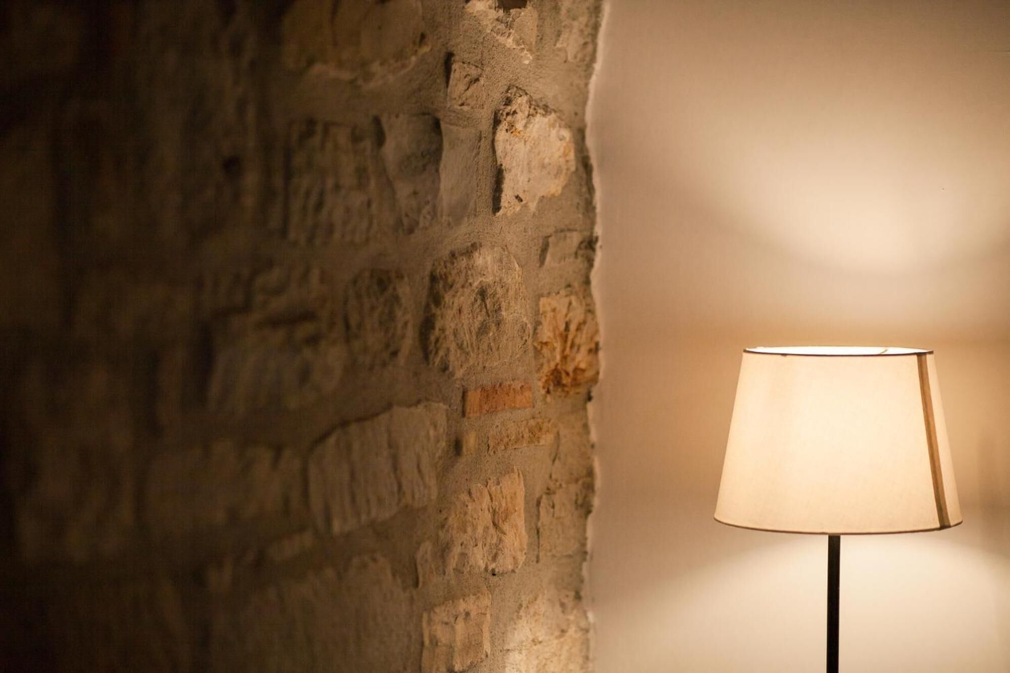 A lamp with a beige shade against a textured stone wall.