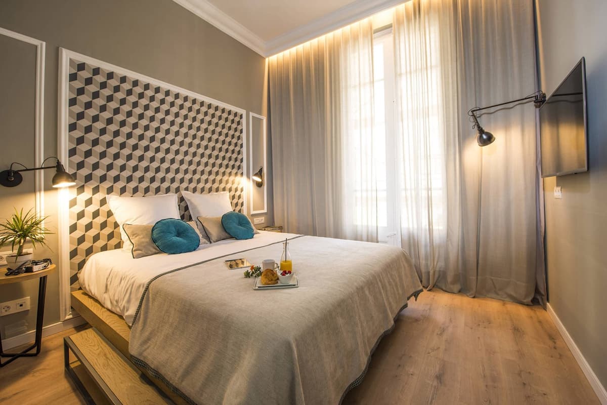 A modern bedroom with a geometric patterned headboard, a bedside table with a lamp, a tray with breakfast items, and a large window with sheer curtains.