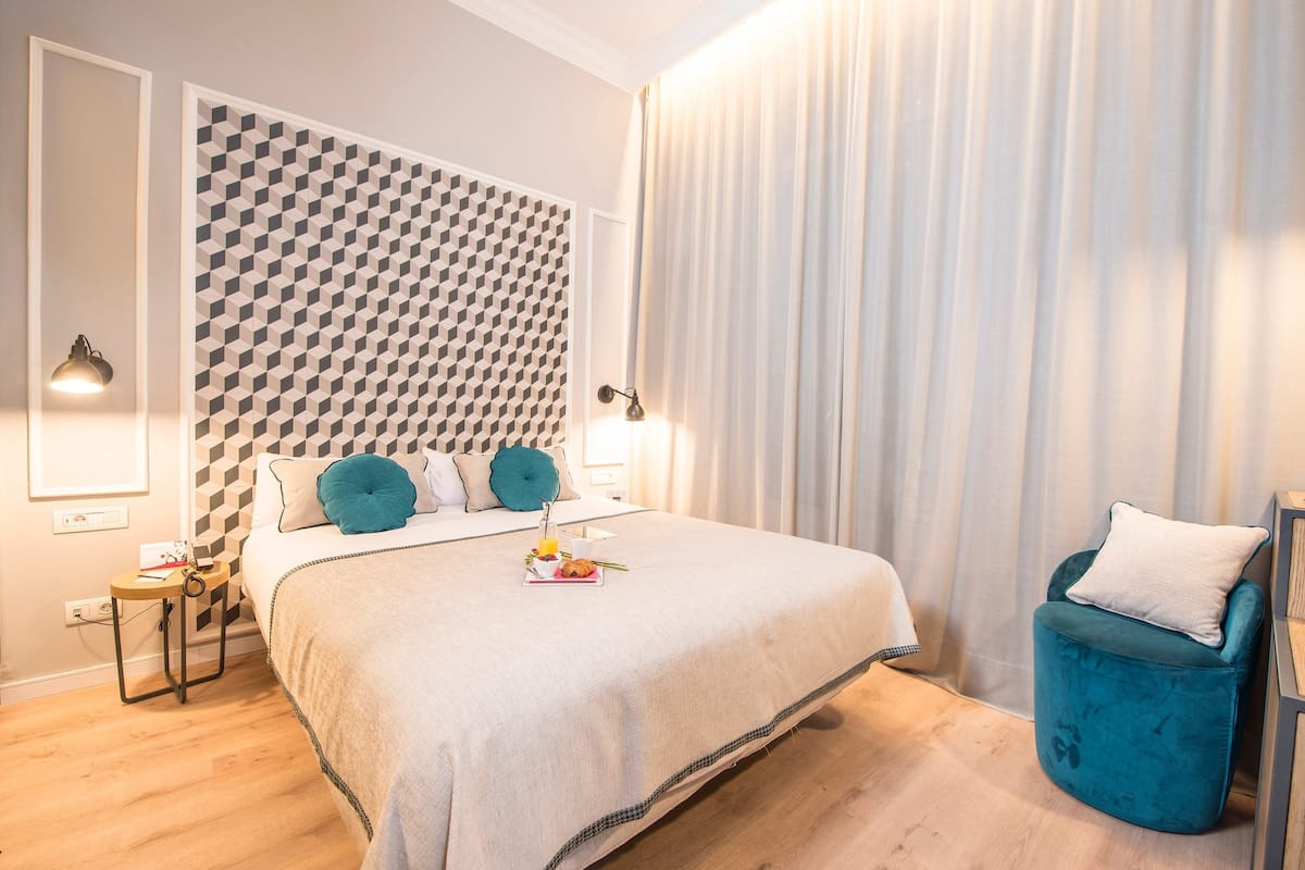A modern bedroom with a geometric-patterned headboard, a bed with a white duvet, teal pillows, and a teal ottoman.