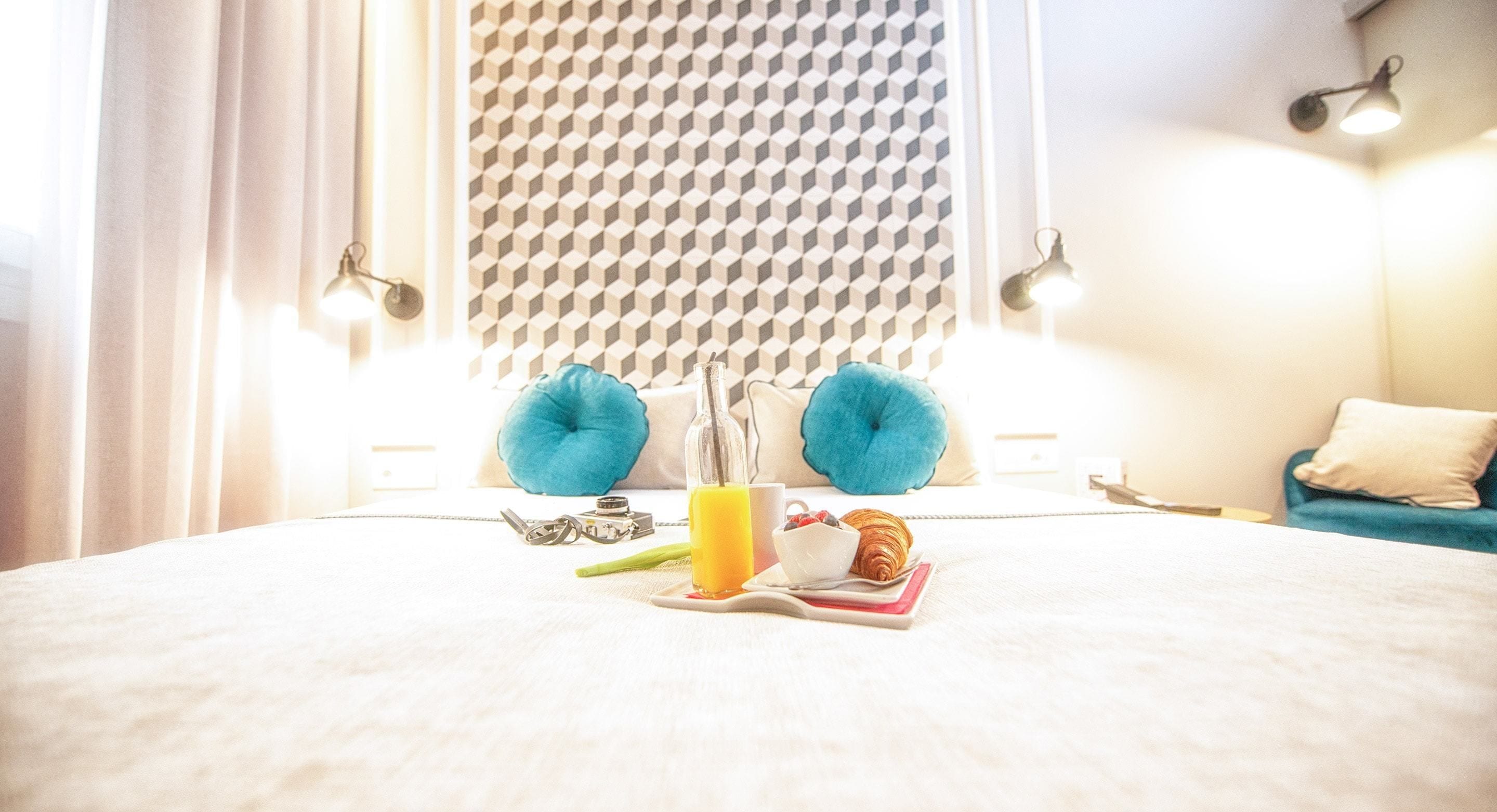A neatly made bed with a bottle of orange juice, a croissant, and a cup, with a blue pillow and a geometric-patterned wall behind.