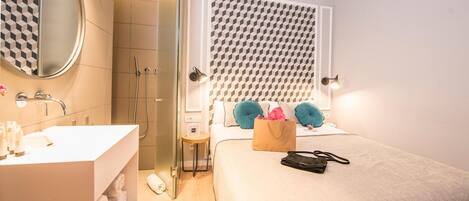 Un baño moderno con un espejo grande, un lavamanos y una ducha. Se observa una cama con cabecera y almohadas estampadas.