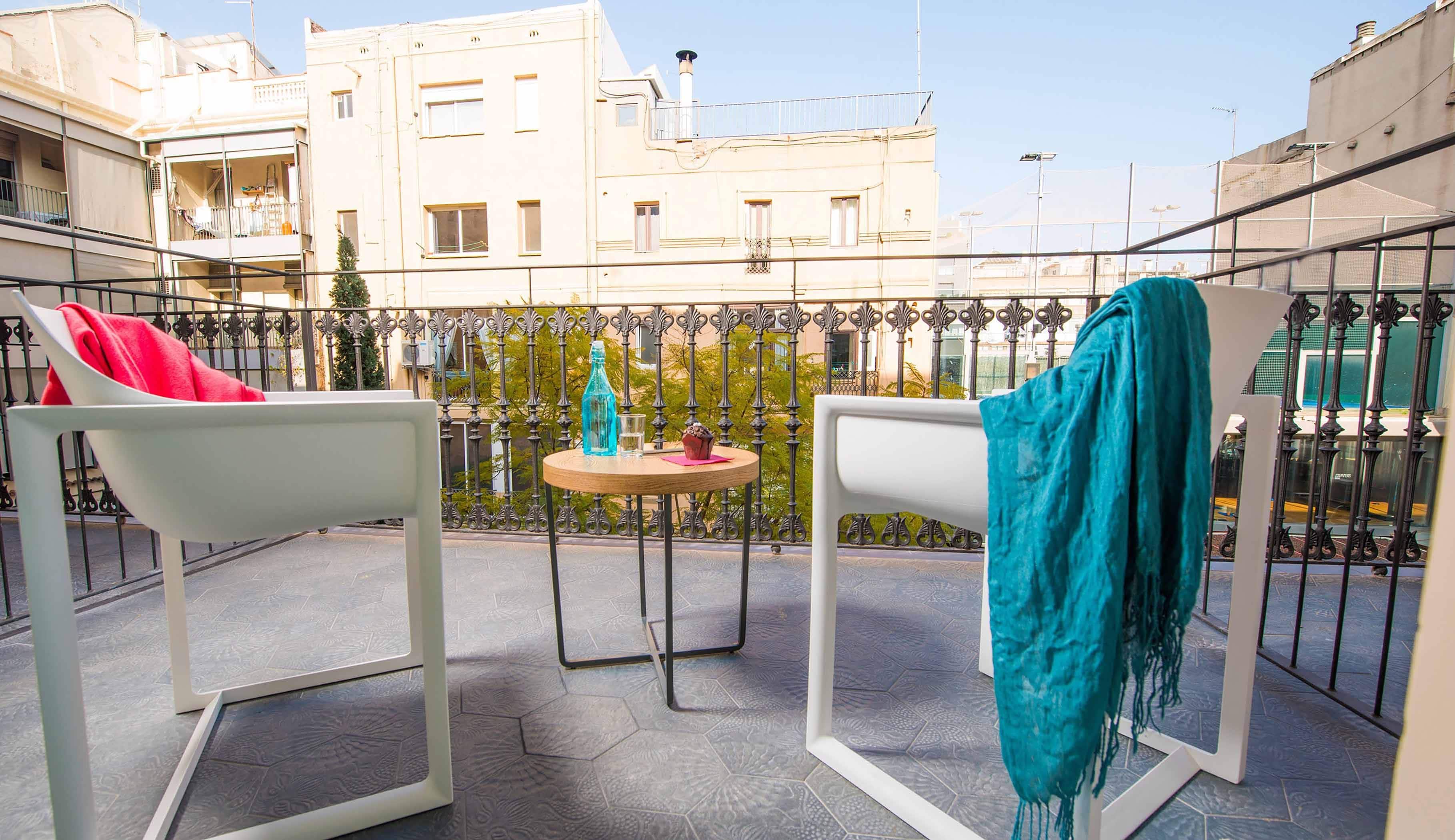 A balcony with two white chairs, one with a red cushion and the other with a teal cushion, a small round table with a bottle and a glass, and a view of buildings in the background.