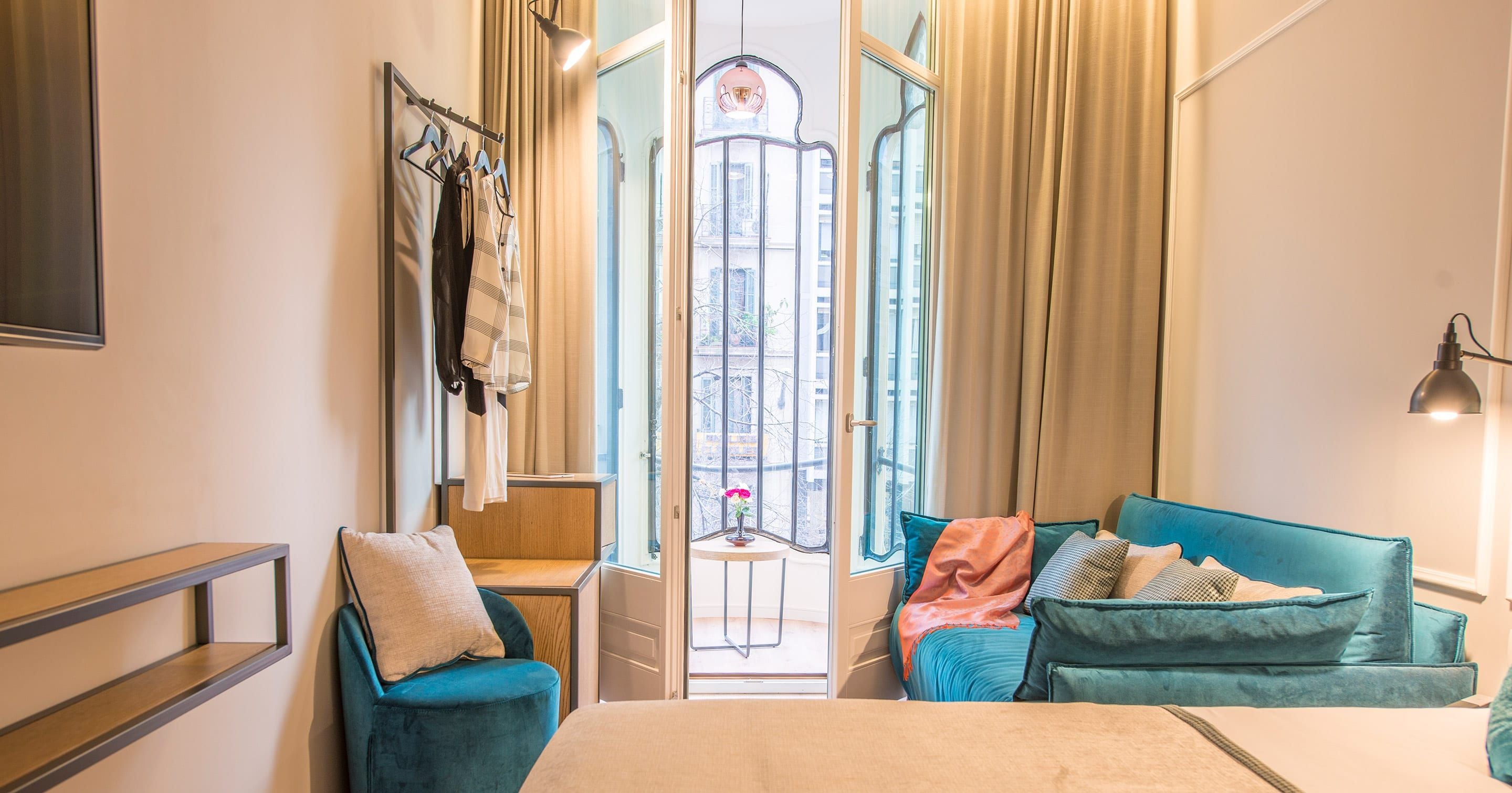 Habitación de hotel con cama, zona de estar con sofá y sillones, ventana con cortinas, lámpara de pared y perchero.