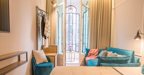 Habitación de hotel con cama, zona de estar con sofá y sillones, ventana con cortinas, lámpara de pared y perchero.