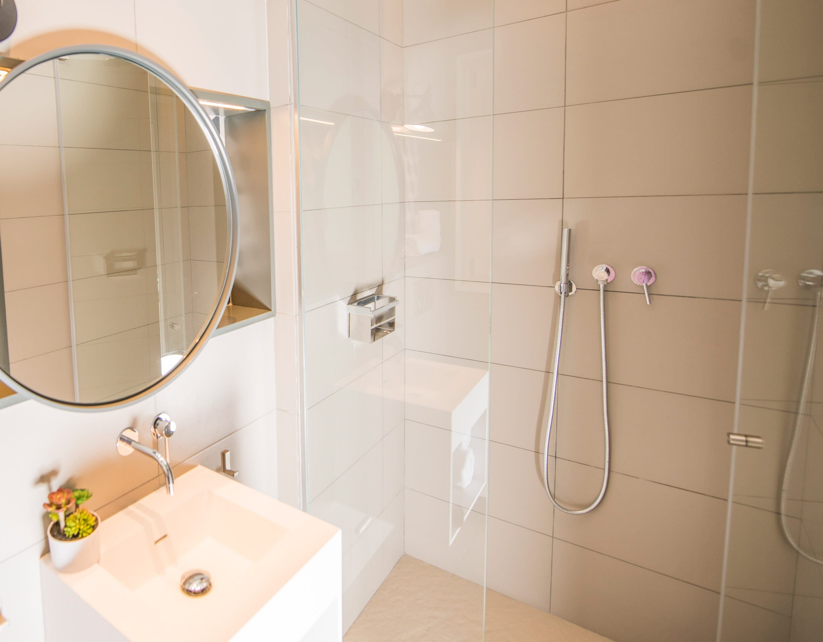 Ein modernes Badezimmer mit rundem Spiegel, Waschbecken und Duschbereich mit zwei Wasserhähnen.