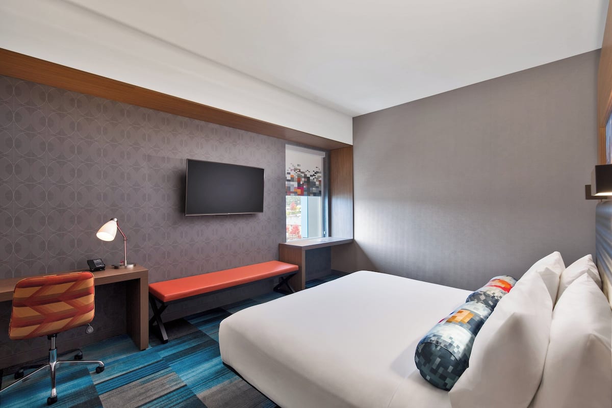 A hotel room with a bed, a desk, a chair, a bench, a TV, and a window.