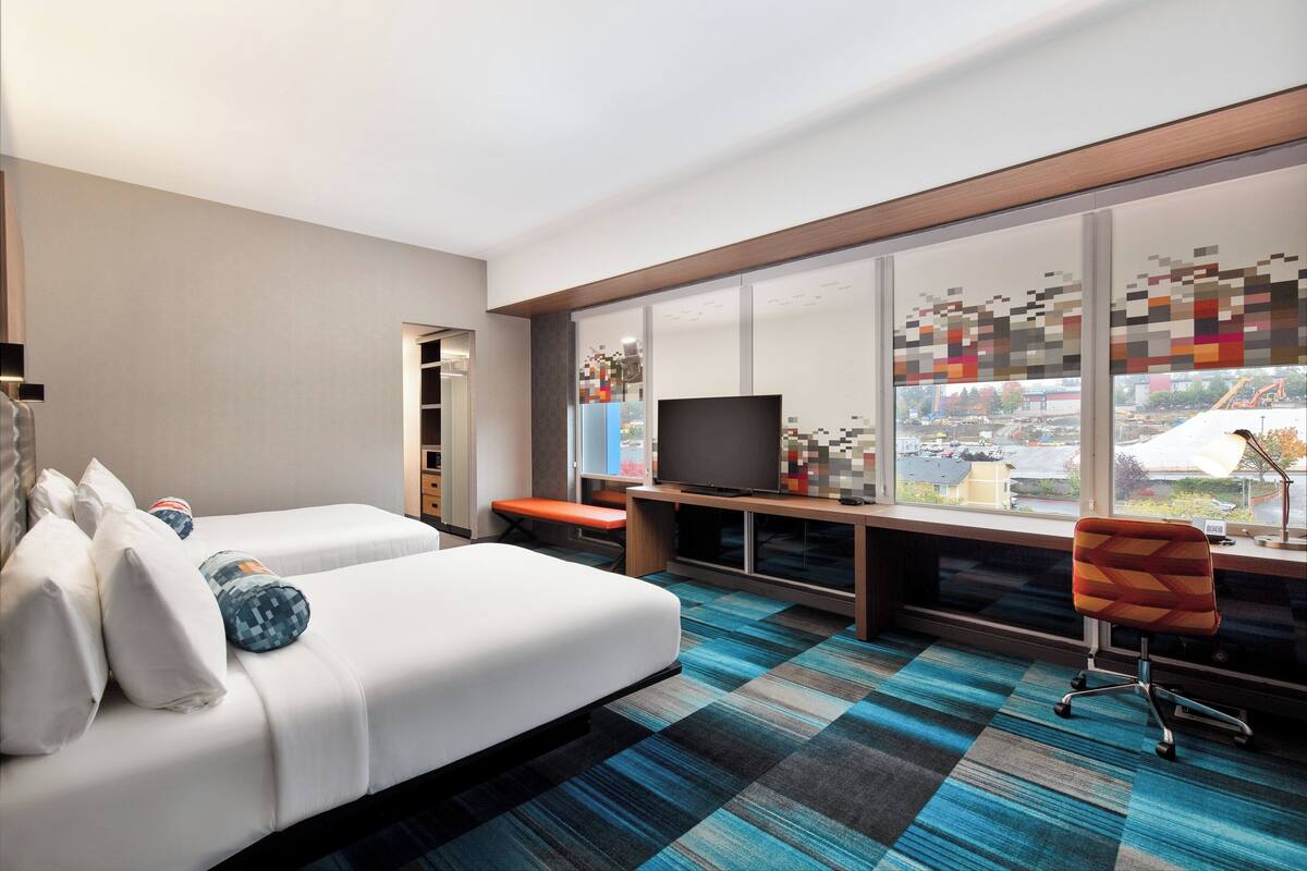 A hotel room with a large window, a bed, a desk with a computer, and a chair.