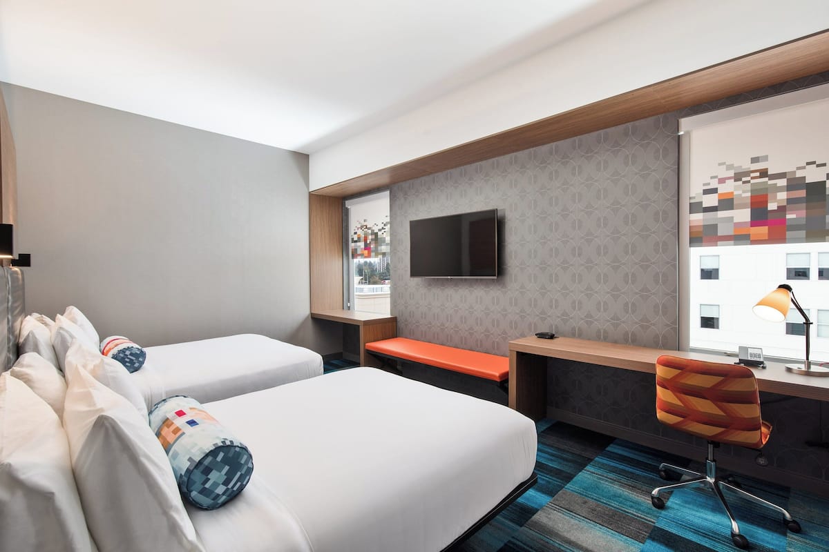 A hotel room with two beds, a desk, a chair, a TV, and a window with a view of buildings.