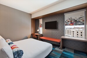 A hotel room with a bed, a desk, a bench, a TV, and a window with a city view. - Aloft by Marriott Seattle Sea-Tac Airport (SeaTac)