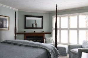 A bedroom with a four-poster bed, a fireplace, a large window with shutters, and a chair.