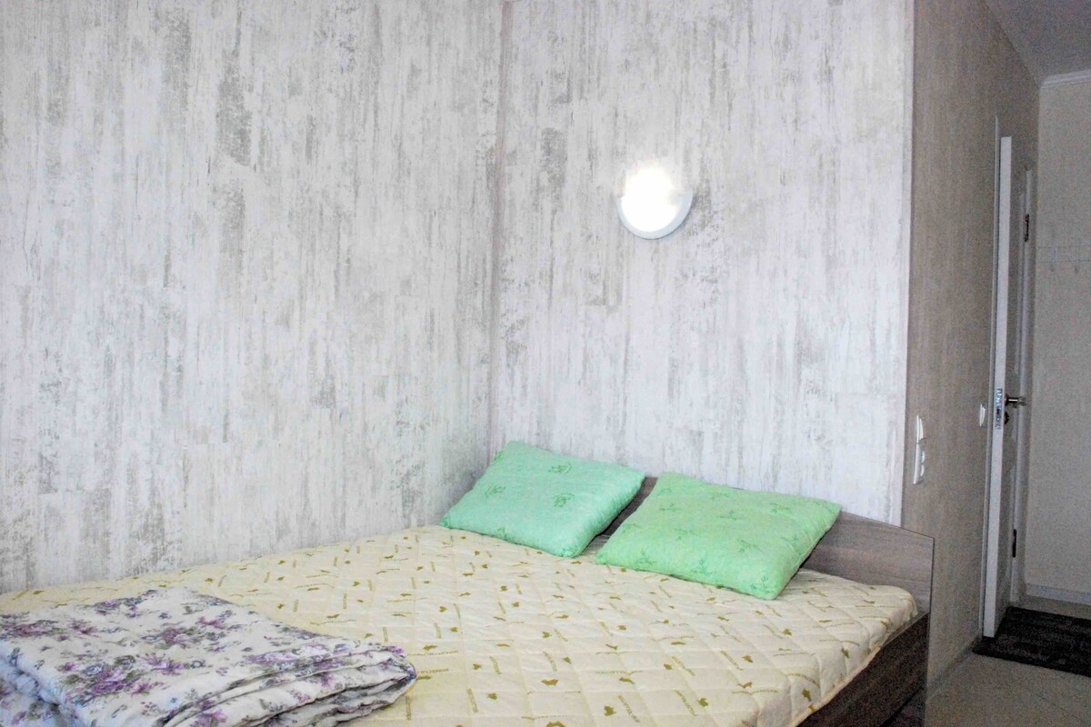 A single bed with a patterned bedspread and two green pillows, a wall-mounted light, and a door to the right.