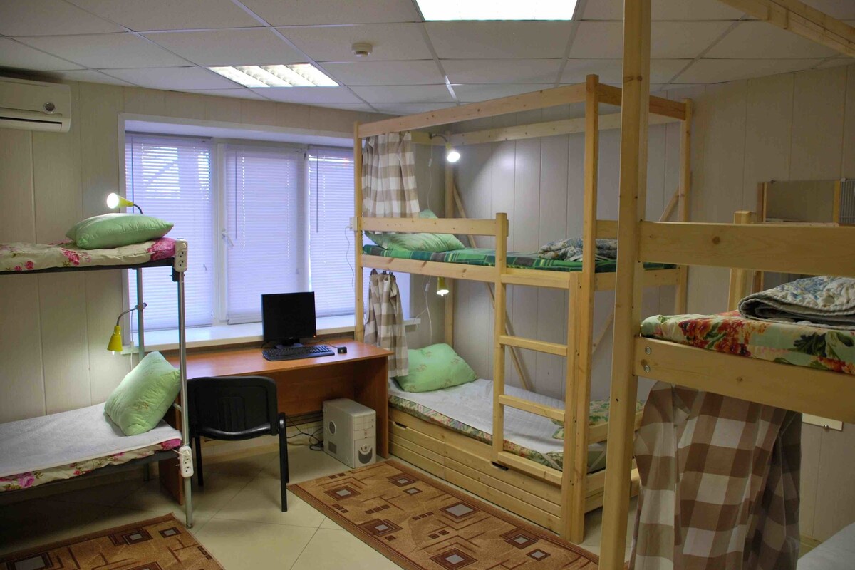 A dormitory room with bunk beds, a desk with a computer, and a window with blinds.