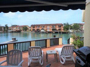 Une terrasse avec deux chaises et un barbecue, donnant sur une étendue d’eau paisible, avec des bâtiments en arrière-plan.