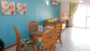 A dining area with wicker chairs and a glass table, a yellow sofa with cushions, and a ceiling fan in a room with blue walls and a large window.