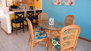 A compact kitchen with wooden cabinets, a microwave, and a coffee maker. There is a dining area with wicker chairs and a glass table.