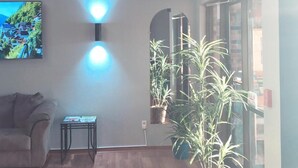 A living room with a sofa, a small table, and a TV mounted on the wall. There are two wall-mounted lights and a large potted plant.